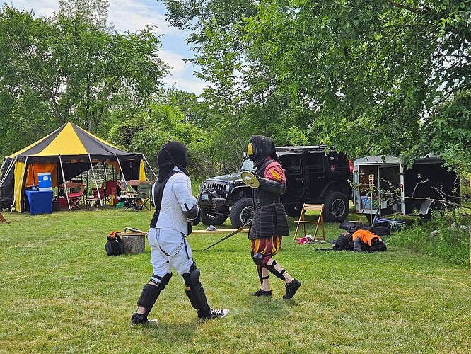 Two people enganged in mixed swordfighting, in full gear. One fighter has a 1-handed sword and shield, and another is fighting with a two-handed sword, not seen int he picture. Behind them is a jeep and a tent.