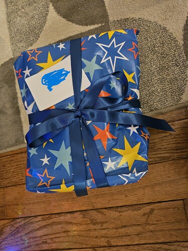 A gift wrapped in wrapping paper with a blue background and white, red, yellow, and lighter blue star pattern. There is a dark blue ribbon forming a bow on the gift.
