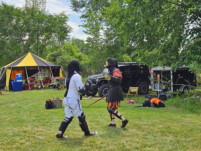 Two people enganged in mixed swordfighting, in full gear. One fighter has a 1-handed sword and shield, and another is fighting with a two-handed sword, not seen int he picture. Behind them is a jeep and a tent.