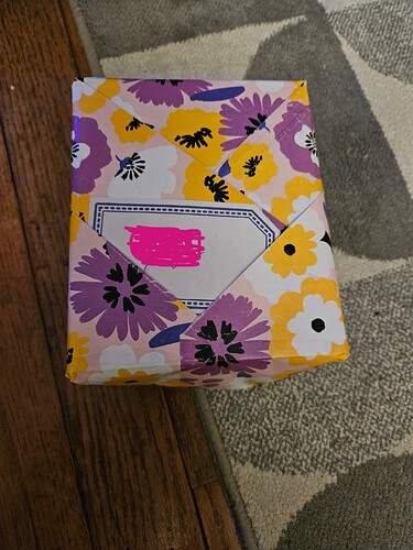A gift wrapped in wrapping paper with a yellow, purple, and white flower pattern. The wrapping paper forms pockets, into one of which the label is inserted.