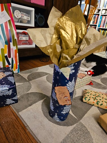 A cylindrical gift wrapped in dark blue wrapping paper patterened with white and light blue Christmas trees, with gold tissue poking from its top.