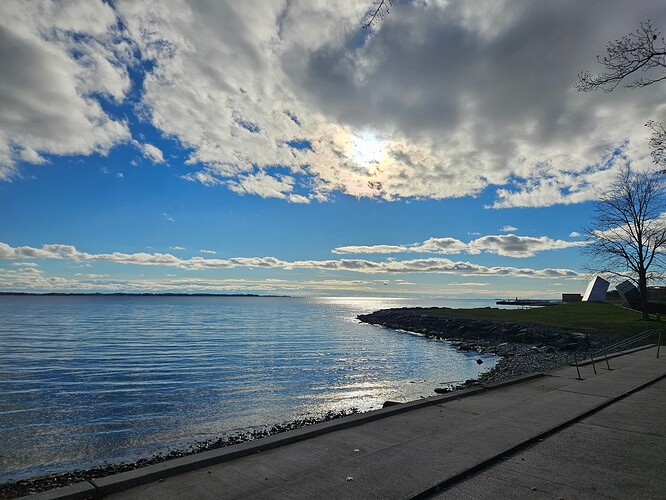 A picture of a calm lake set against a bright blue sky with white clouds. To the right of the picture, a metal sculpture and some trees can be seen in the distance.