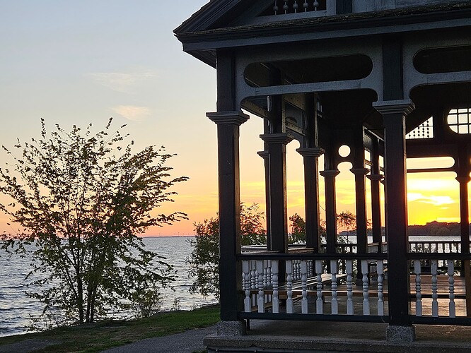 A gazebo is partially seen ont he right, with a tree and a lake to the left, and the sunset in the background.