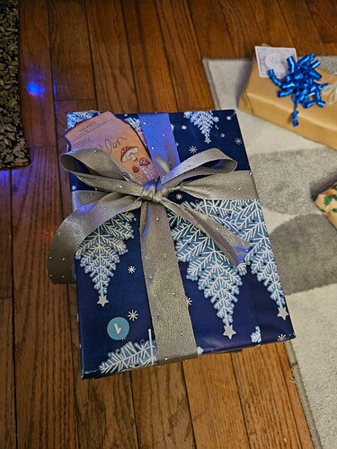 A gift wrapped in blue wrapping paper with white and blue Christmas trees, and a silver bow with small, white, raised dots.