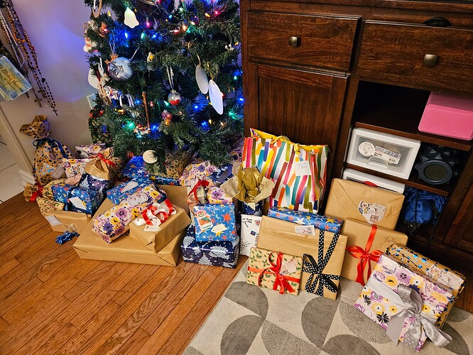 Several presents wrapped in bows of various colours, under a decorated Christmas tree on a hardwood floor on the left, with a patterned rug on the right.