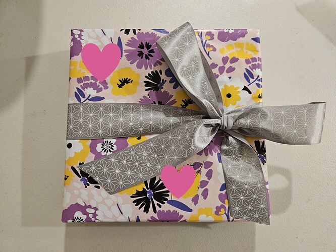 A gift wrapped in wrapping paper with a purple, yellow, and white flower pattern. The gift is wrapped with a silver ribbon with a white star pattern, which has been formed into a bow. Pink hearts have been added digitally to hide the recipient's name.