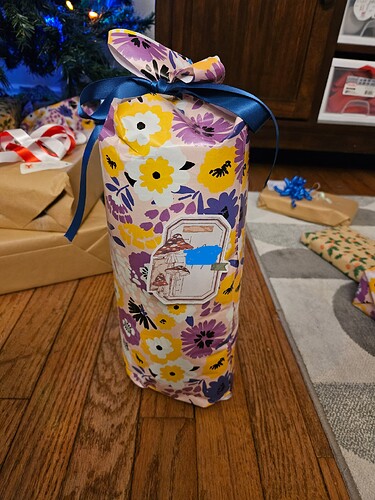 A gift wrapped in wrapping paper with a yellow, purple, and white flower pattern, in a cylindrical shape, with a blue bow at the top.